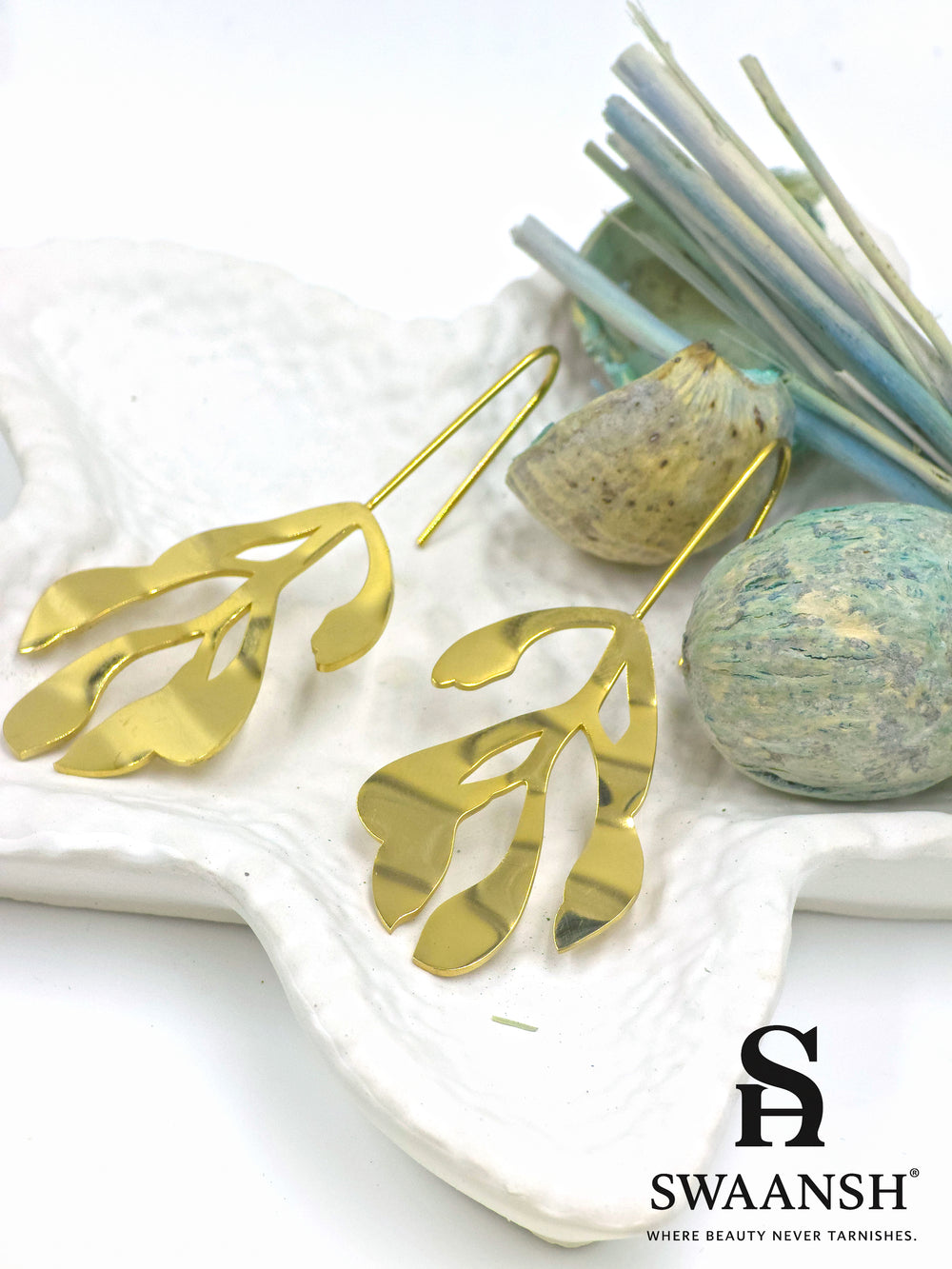 Leaf Muse gold sculptural earrings in open leaf design with artistic hook-back by Swaansh, styled on a white plate with beige flowers