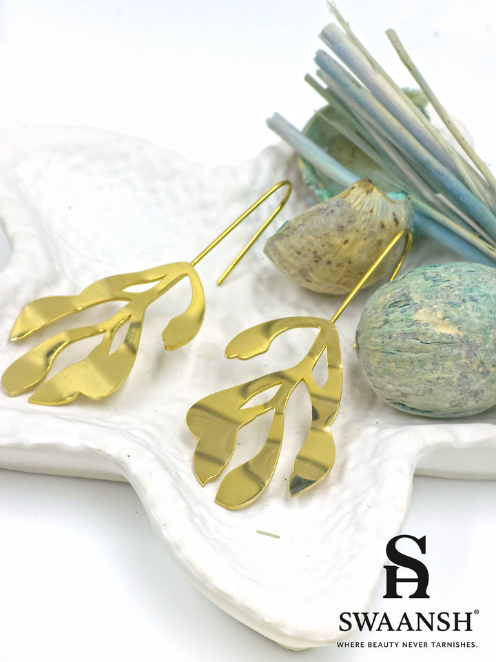 Leaf Muse gold sculptural earrings in open leaf design with artistic hook-back by Swaansh, styled on a white plate with beige flowers