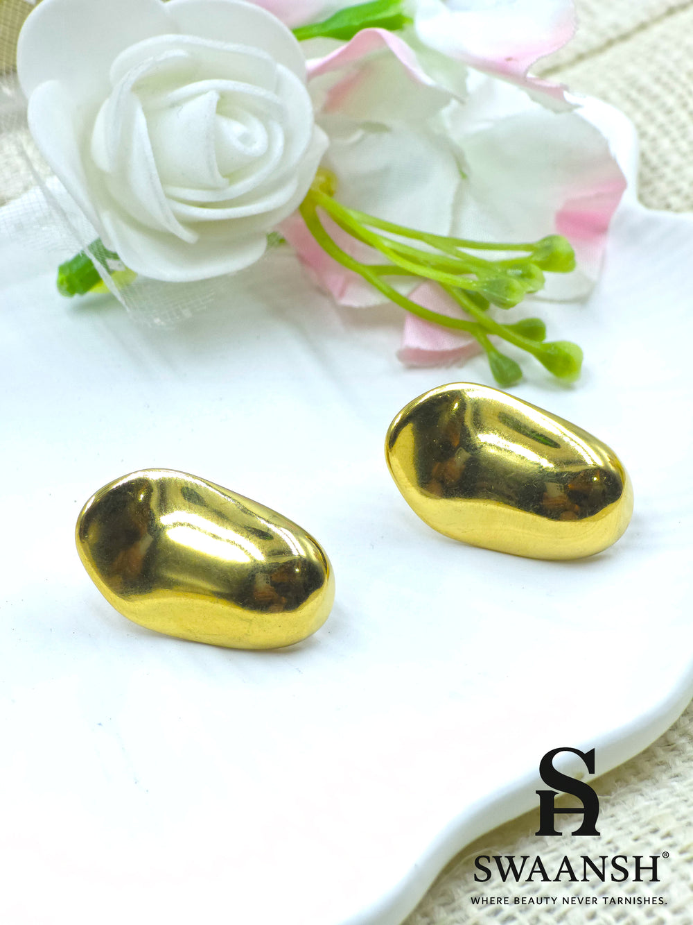 Swaansh Molten Muse earrings – glossy nugget-shaped sculptural gold studs placed on white textured background with dried blue botanicals