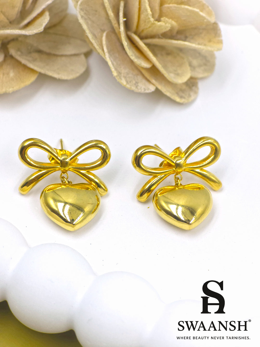 PuffHeart Bows – polished gold earrings with heart drops and knot bows styled on a white ceramic plate with floral backdrop