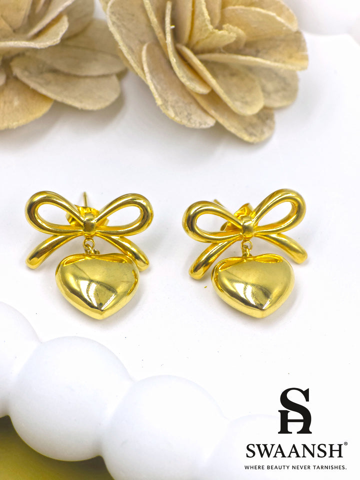 PuffHeart Bows – polished gold earrings with heart drops and knot bows styled on a white ceramic plate with floral backdrop