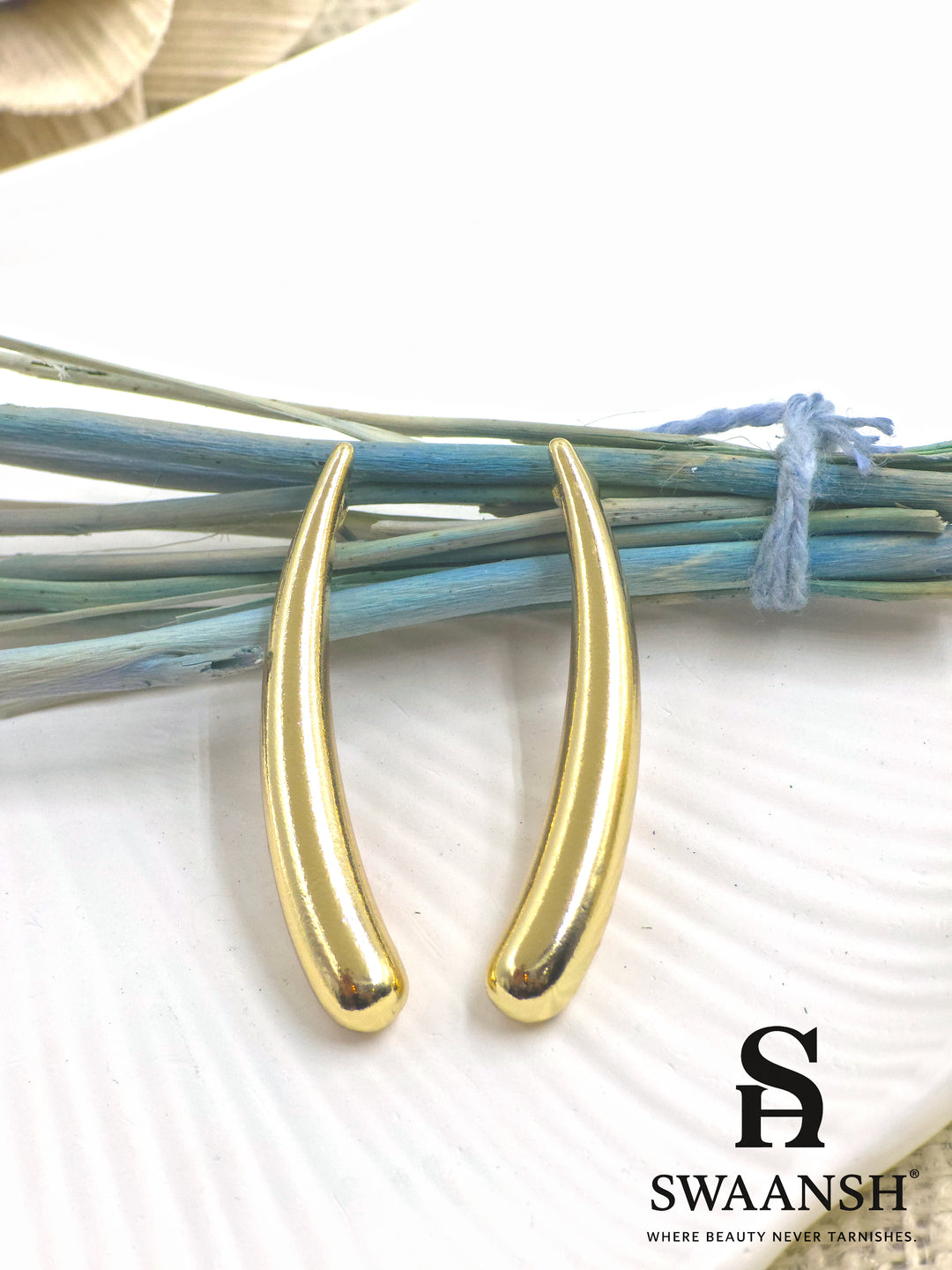 SwayDrop sleek curved gold earrings placed on a white ceramic plate with dried blue floral accents – premium anti-tarnish jewellery by Swaansh