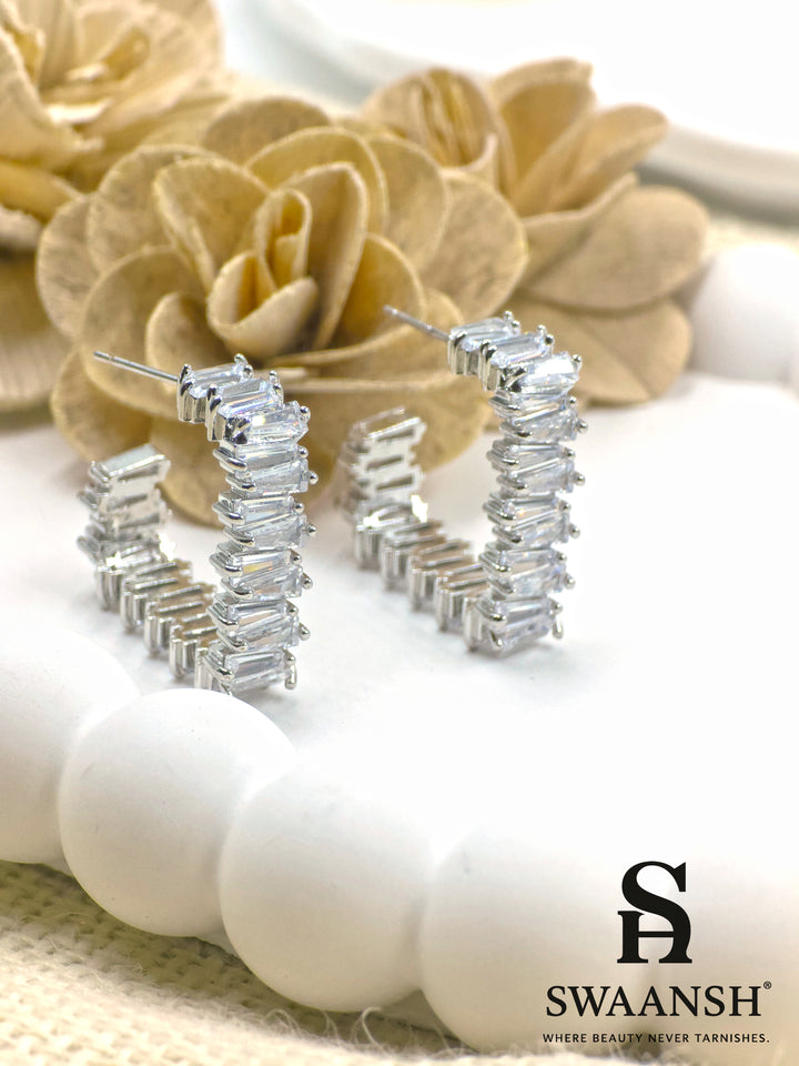 Silver semi-hoop earrings with baguette-cut stones placed on a luxury floral setup – Swaansh