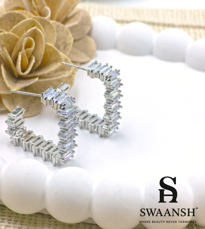 Silver semi-hoop earrings with baguette-cut stones placed on a luxury floral setup – Swaansh