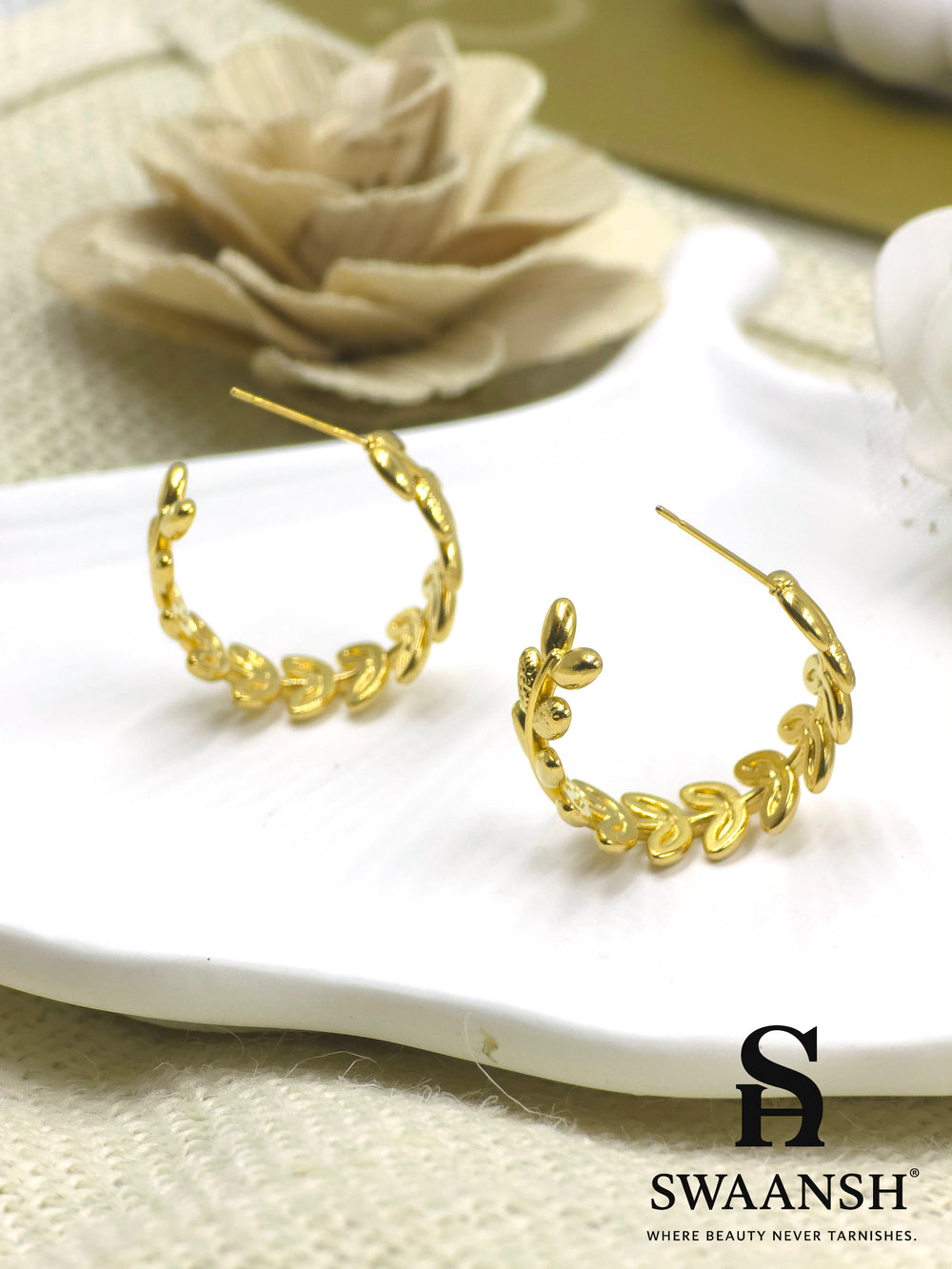 Swaansh Golden Laurel earrings – gold hoop earrings with delicate laurel leaf design, placed on a white plate with floral background