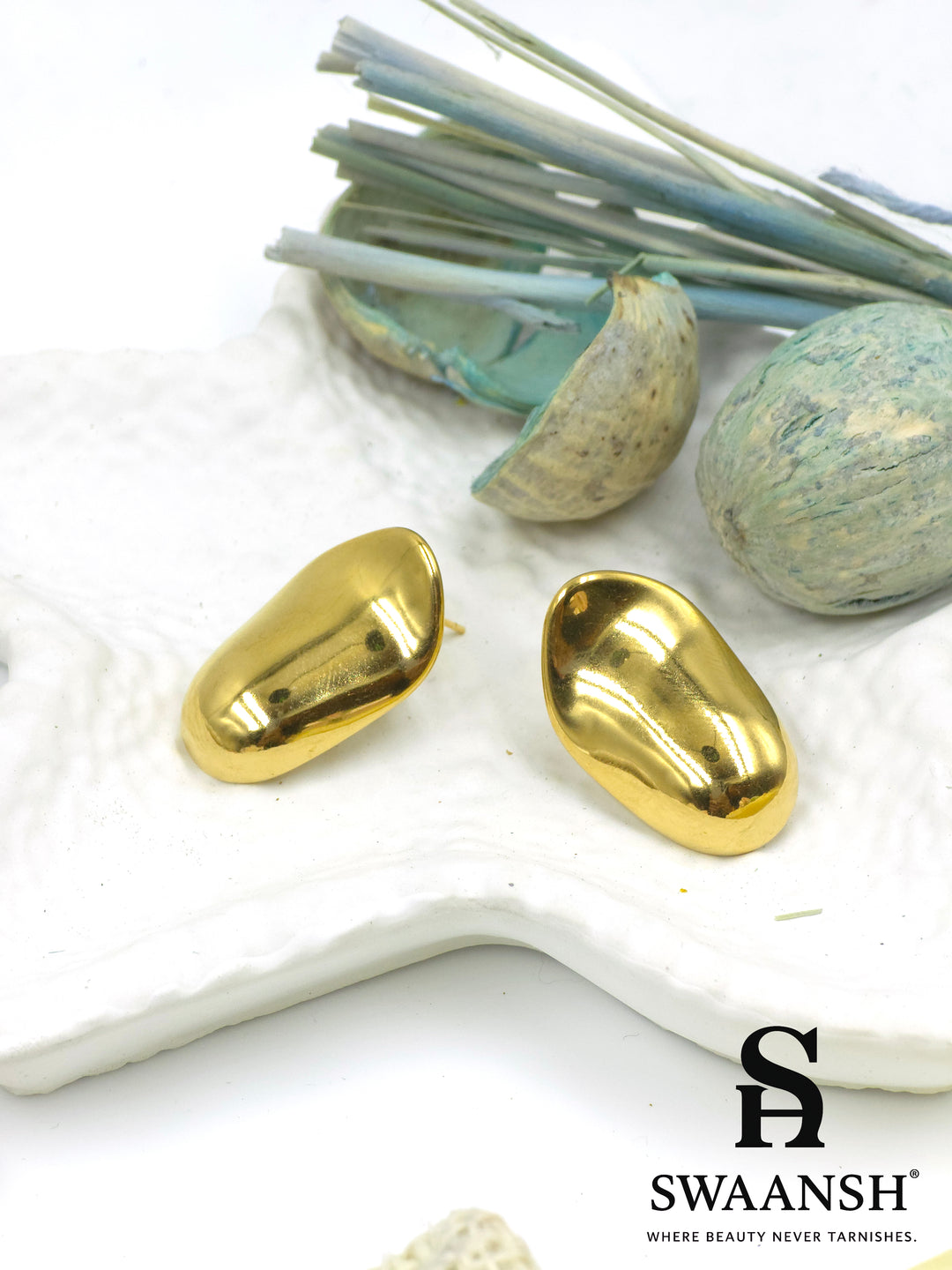 Swaansh Molten Muse earrings – glossy nugget-shaped sculptural gold studs placed on white textured background with dried blue botanicals