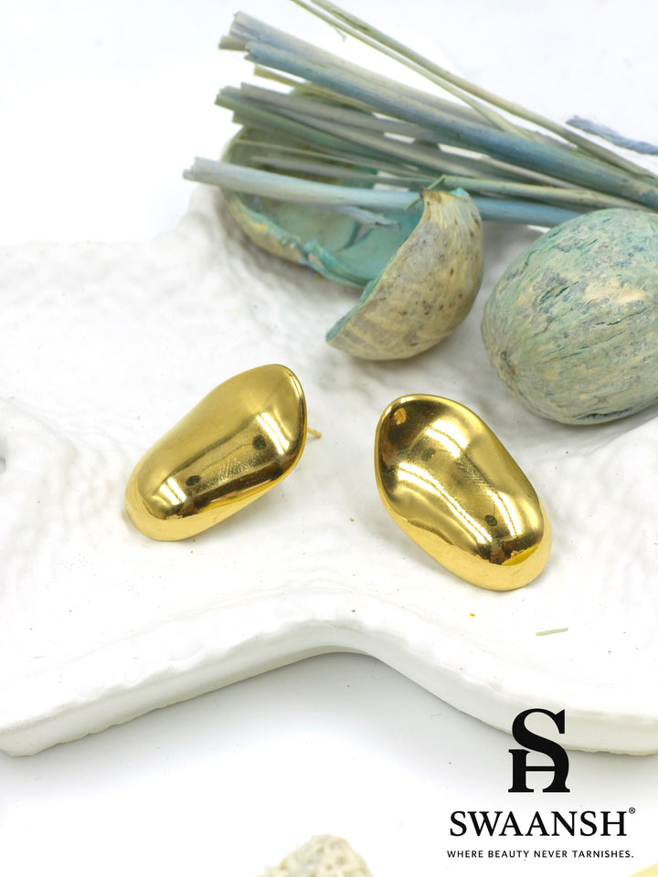 Swaansh Molten Muse earrings – glossy nugget-shaped sculptural gold studs placed on white textured background with dried blue botanicals