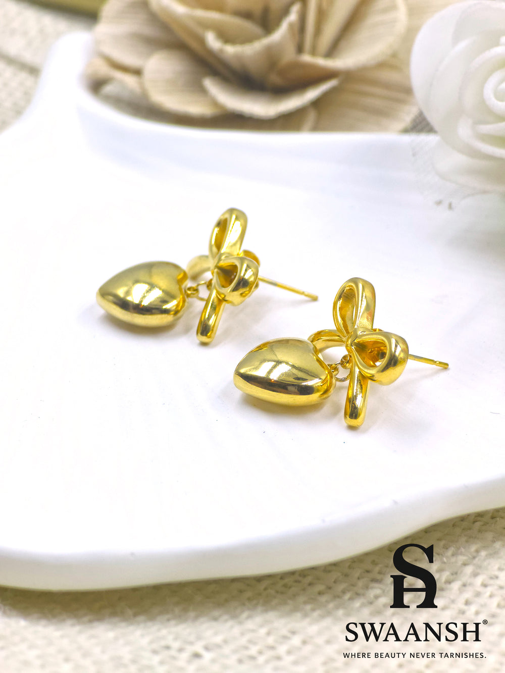PuffHeart Bows – polished gold earrings with heart drops and knot bows styled on a white ceramic plate with floral backdrop