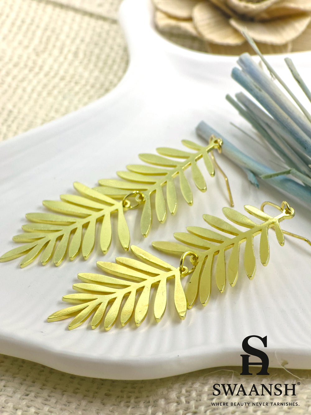 PalmWhisper – tropical palm leaf gold earrings styled on a white ceramic plate with dried flowers in background