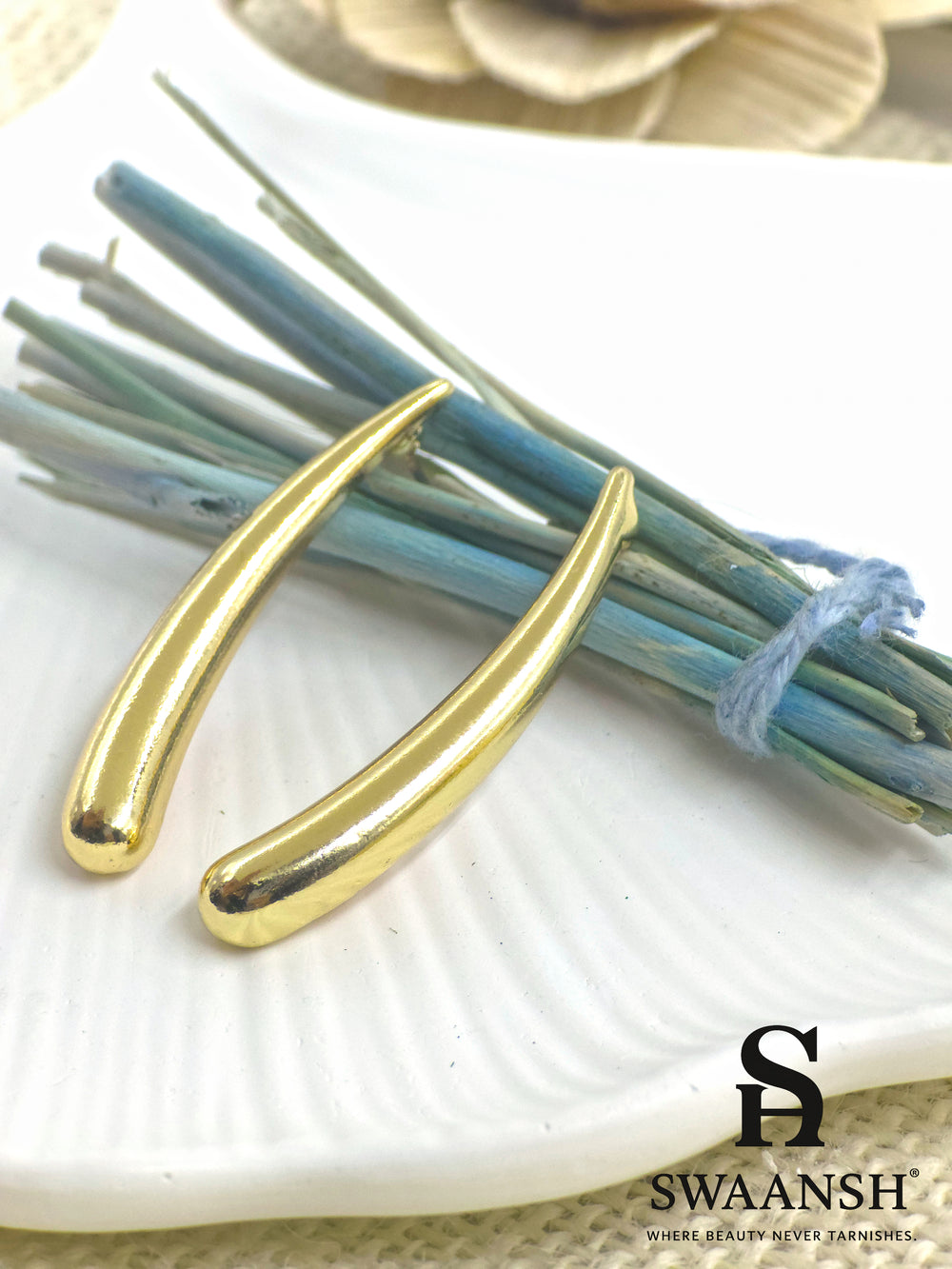 SwayDrop sleek curved gold earrings placed on a white ceramic plate with dried blue floral accents – premium anti-tarnish jewellery by Swaansh