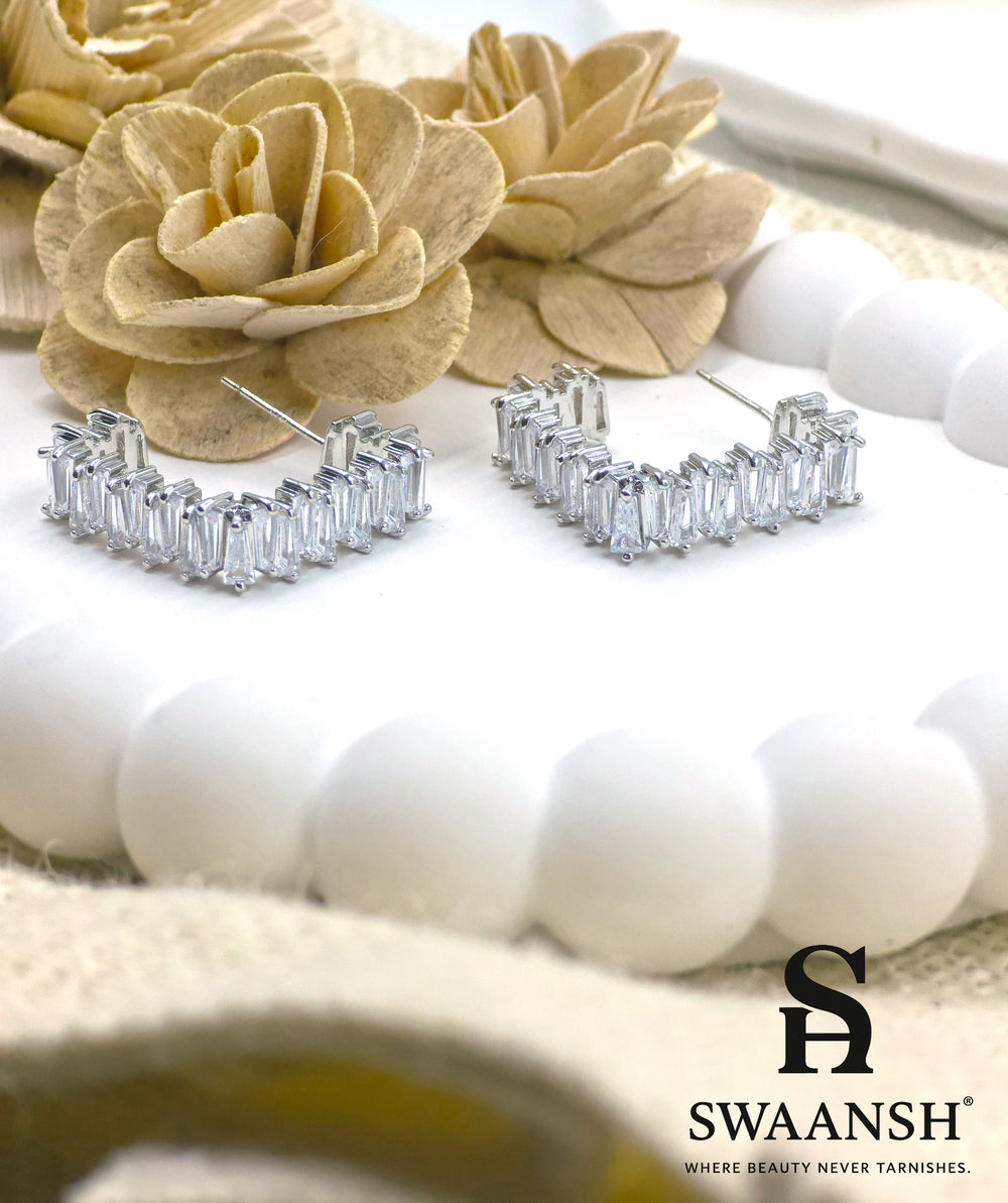Silver semi-hoop earrings with baguette-cut stones placed on a luxury floral setup – Swaansh